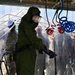 A military personnel wearing full CBRN (Chemical, Biological, Radiological, and Nuclear) protective gear, including a gas mask and olive-green hazmat suit with black rubber gloves, is undergoing a decontamination procedure inside a transparent plastic curtain enclosure. Red and blue coiled hoses are suspended overhead, likely part of a decontamination wash system. Several other military personnel in standard camouflage uniforms observe from outside the enclosure.