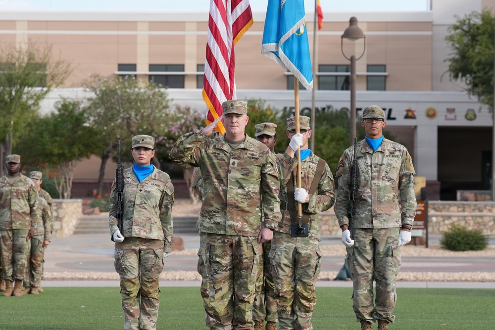 A U.S. Army military ceremony is taking place on a parade field, likely at a stateside installation. A central officer is rendering a salute while color guard members hold the American flag and a light blue unit guidon. Additional soldiers in formation are visible in the background wearing OCP (Operational Camouflage Pattern) uniforms.