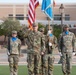 A U.S. Army military ceremony is taking place on a parade field, likely at a stateside installation. A central officer is rendering a salute while color guard members hold the American flag and a light blue unit guidon. Additional soldiers in formation are visible in the background wearing OCP (Operational Camouflage Pattern) uniforms.