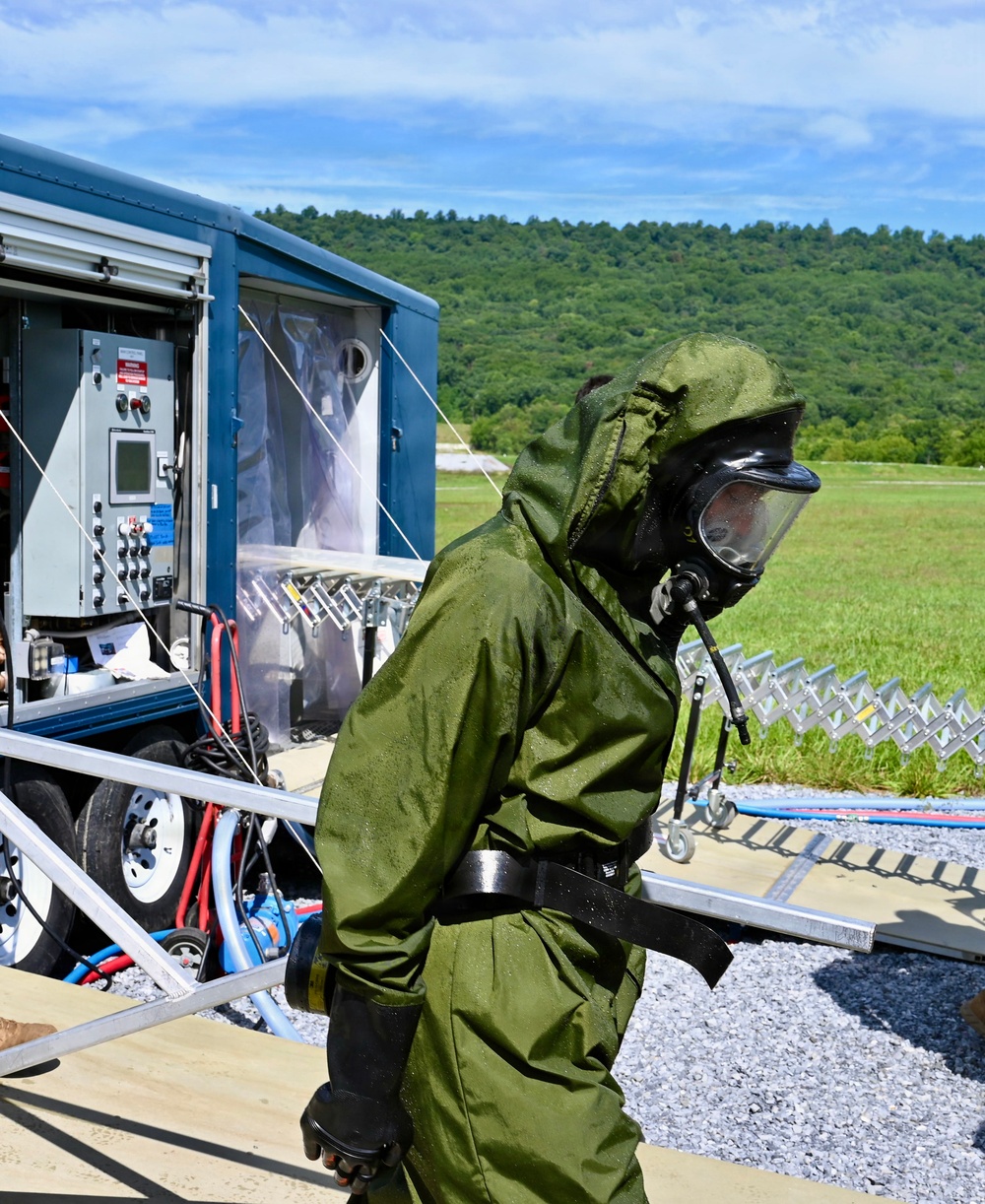 A person wearing a full olive-green CBRN (Chemical, Biological, Radiological, Nuclear) protective suit and gas mask is visible in the foreground, appearing wet from decontamination procedures. Behind them is a blue mobile decontamination trailer unit with control panels, hoses, and plastic curtain barriers. The setting appears to be an outdoor training or operational exercise area with green fields and forested hills in the background.