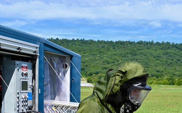 A person wearing a full olive-green CBRN (Chemical, Biological, Radiological, Nuclear) protective suit and gas mask is visible in the foreground, appearing wet from decontamination procedures. Behind them is a blue mobile decontamination trailer unit with control panels, hoses, and plastic curtain barriers. The setting appears to be an outdoor training or operational exercise area with green fields and forested hills in the background.