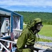 A person wearing a full olive-green CBRN (Chemical, Biological, Radiological, Nuclear) protective suit and gas mask is visible in the foreground, appearing wet from decontamination procedures. Behind them is a blue mobile decontamination trailer unit with control panels, hoses, and plastic curtain barriers. The setting appears to be an outdoor training or operational exercise area with green fields and forested hills in the background.