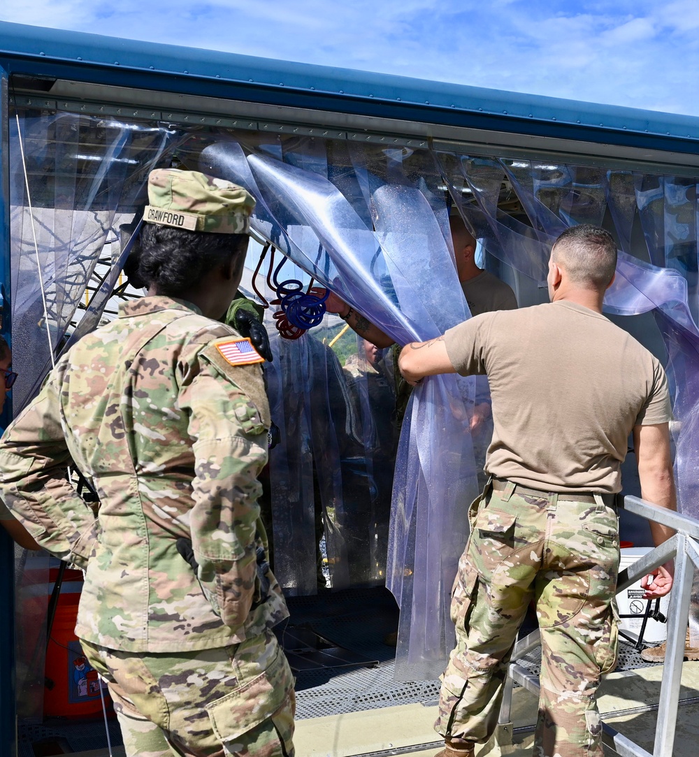U.S. Army soldiers are working with what appears to be a decontamination or CBRN (Chemical, Biological, Radiological, Nuclear) station, characterized by clear plastic strip curtains forming an enclosed passageway. At least three soldiers in OCP (Operational Camouflage Pattern) uniforms are visible, with one identified by a name tape reading 'CRAWFORD.' They appear to be setting up, adjusting, or operating the portable decontamination unit mounted on a trailer or platform.