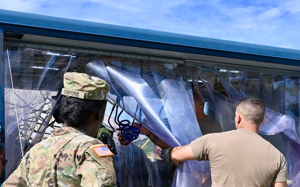 U.S. Army soldiers are working with what appears to be a decontamination or CBRN (Chemical, Biological, Radiological, Nuclear) station, characterized by clear plastic strip curtains forming an enclosed passageway. At least three soldiers in OCP (Operational Camouflage Pattern) uniforms are visible, with one identified by a name tape reading 'CRAWFORD.' They appear to be setting up, adjusting, or operating the portable decontamination unit mounted on a trailer or platform.