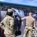 U.S. Army soldiers are working with what appears to be a decontamination or CBRN (Chemical, Biological, Radiological, Nuclear) station, characterized by clear plastic strip curtains forming an enclosed passageway. At least three soldiers in OCP (Operational Camouflage Pattern) uniforms are visible, with one identified by a name tape reading 'CRAWFORD.' They appear to be setting up, adjusting, or operating the portable decontamination unit mounted on a trailer or platform.