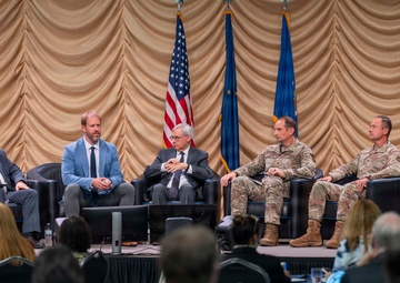 A formal panel discussion or symposium is taking place on a stage before a large seated audience. Five panelists — three in civilian business attire and two in military camouflage uniforms — are seated in black leather chairs on a raised platform. The backdrop features draped beige curtains and three flags, including the United States flag and what appear to be U.S. Air Force flags.
