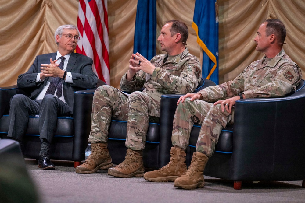 Three individuals are seated in black leather chairs on a stage during what appears to be a formal panel discussion or symposium. Two individuals are wearing U.S. Air Force camouflage uniforms (OCP pattern), while one is dressed in civilian business attire. An American flag and a U.S. Air Force flag are visible in the background.