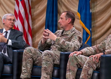 Three individuals are seated in black leather chairs on a stage during what appears to be a formal panel discussion or symposium. Two individuals are wearing U.S. Air Force camouflage uniforms (OCP pattern), while one is dressed in civilian business attire. An American flag and a U.S. Air Force flag are visible in the background.
