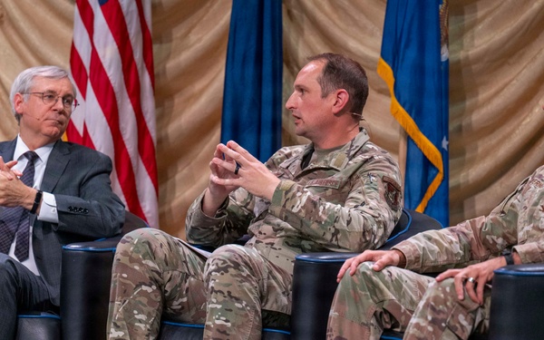 Three individuals are seated in black leather chairs on a stage during what appears to be a formal panel discussion or symposium. Two individuals are wearing U.S. Air Force camouflage uniforms (OCP pattern), while one is dressed in civilian business attire. An American flag and a U.S. Air Force flag are visible in the background.