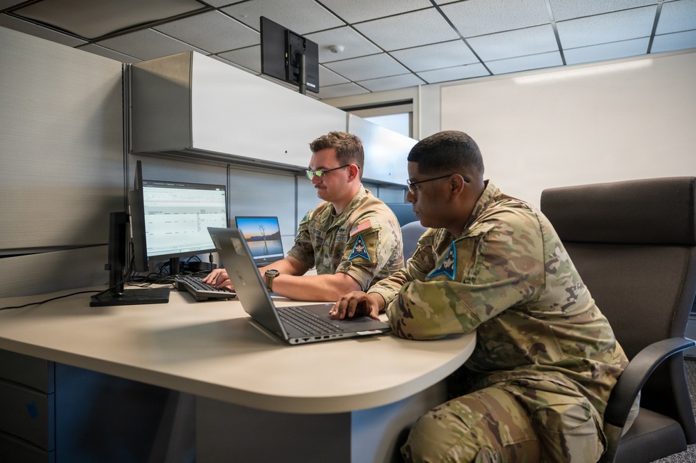 Two military personnel in OCP (Operational Camouflage Pattern) uniforms are working together at a modern office workstation. They appear to be reviewing data on both a laptop and an external monitor, likely in a military administrative or operations facility. Both individuals wear U.S. flag patches and unit insignia consistent with U.S. Space Force or U.S. Air Force.