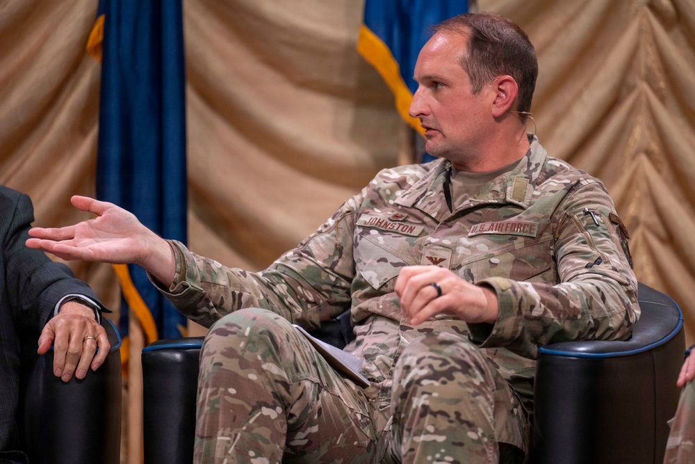 A U.S. Air Force officer with the name tape 'JOHNSTON' is seated in a black chair, gesturing with his right hand while speaking at what appears to be a panel discussion or formal event. He is wearing OCP (Operational Camouflage Pattern) uniform with a colonel's eagle insignia visible on his chest. The background features draped gold and blue curtains along with what appears to be a U.S. Air Force flag.