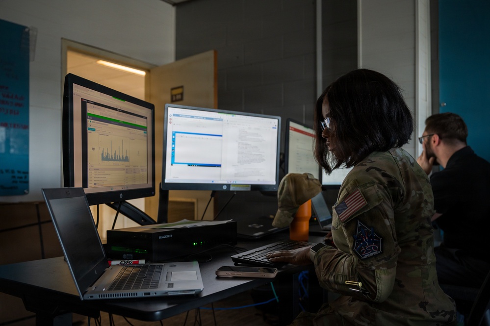 A U.S. military service member in camouflage uniform is working at a multi-monitor computer workstation, appearing to analyze data or conduct cyber/IT operations. The setting appears to be a military operations or cyber operations center. A second person in civilian attire is visible in the background also working at a computer.