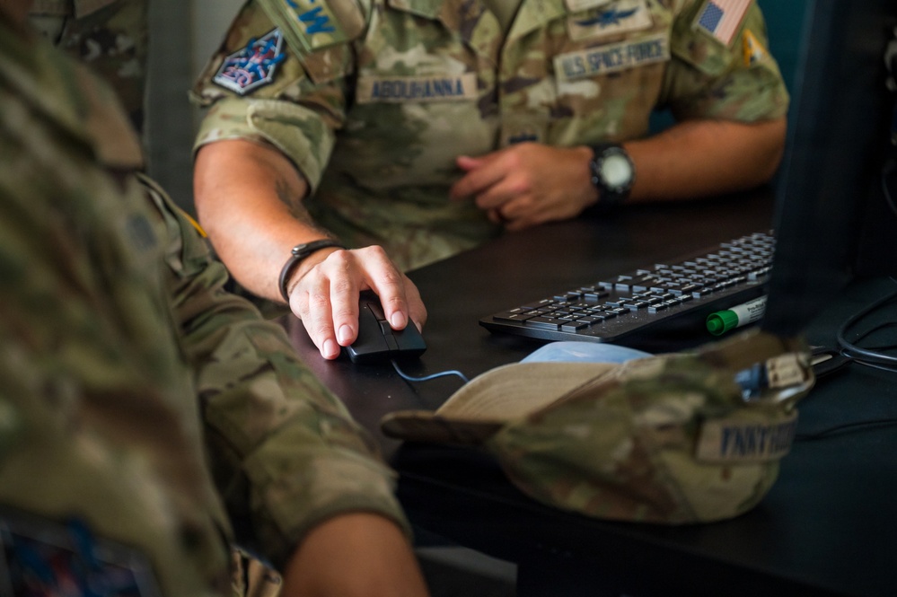 Military personnel in OCP (Operational Camouflage Pattern) uniforms are working at a computer workstation. At least one individual is identifiable as U.S. Space Force based on the visible branch tape reading 'U.S. SPACE FORCE' and associated patches. The scene depicts a collaborative work environment with personnel gathered around a desk with a keyboard and mouse.