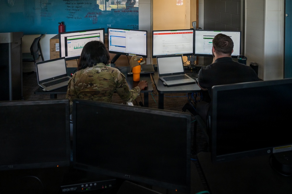 Two individuals are seated at workstations in what appears to be a military or government operations center, working on multiple monitors and laptops. One person on the left is wearing military camouflage (OCP/MultiCam) uniform, while the person on the right is in civilian dark clothing. The workspace features multiple large monitors displaying various software interfaces, laptops, and a whiteboard with writing on the wall.