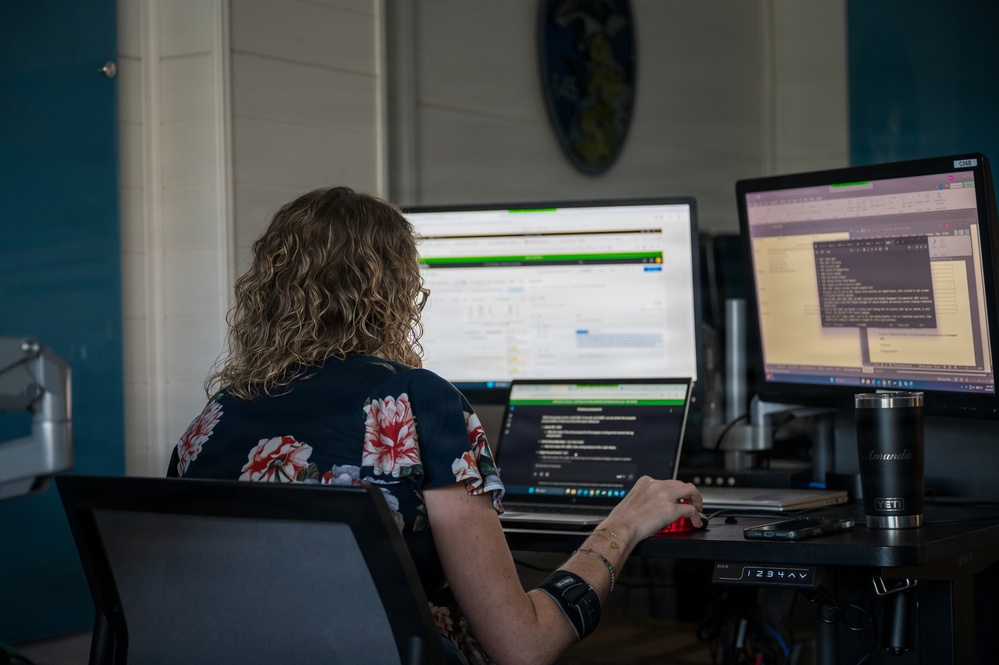 A person with curly blonde hair, wearing a dark floral-print top, is seated at a multi-monitor workstation viewed from behind. The setup includes a laptop, two external monitors displaying what appears to be software development or data management interfaces, and a YETI tumbler on the desk. The workspace is in a room with teal/blue walls and white shiplap paneling.