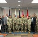 A group photo of approximately 20 individuals, a mix of U.S. Army military personnel in OCP (Operational Camouflage Pattern) uniforms and civilians in business casual attire, posing together in what appears to be a military conference or briefing room. Multiple military flags are displayed in the background, including the U.S. flag and what appear to be Army unit colors. The setting suggests an official military ceremony, meeting, or recognition event.
