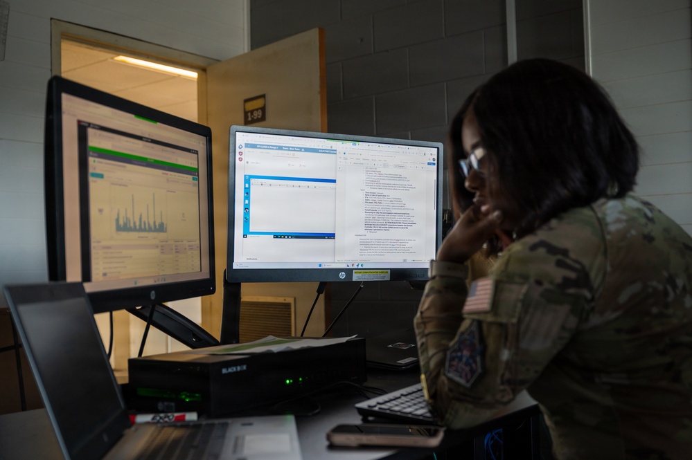 A military service member in camouflage uniform (OCP/MultiCam pattern) with a US flag patch is working at a dual-monitor computer workstation in what appears to be a military office or operations center. The individual is reviewing documents or data displayed across two HP monitors while in a contemplative pose with hand raised to chin. The workspace includes a Black Box network device and a laptop.