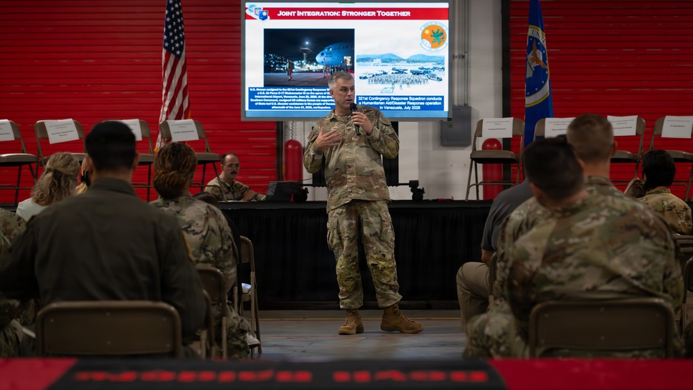 A military officer in camouflage uniform is delivering a briefing or presentation to an audience of seated military personnel in what appears to be a hangar or large facility. The presentation slide behind him is titled 'Joint Integration: Stronger Together' and references the 321st Contingency Response Squadron conducting a Humanitarian Aid/Disaster Response operation in Venezuela, July 2026. An American flag and what appears to be a unit flag are visible flanking the presentation screen.