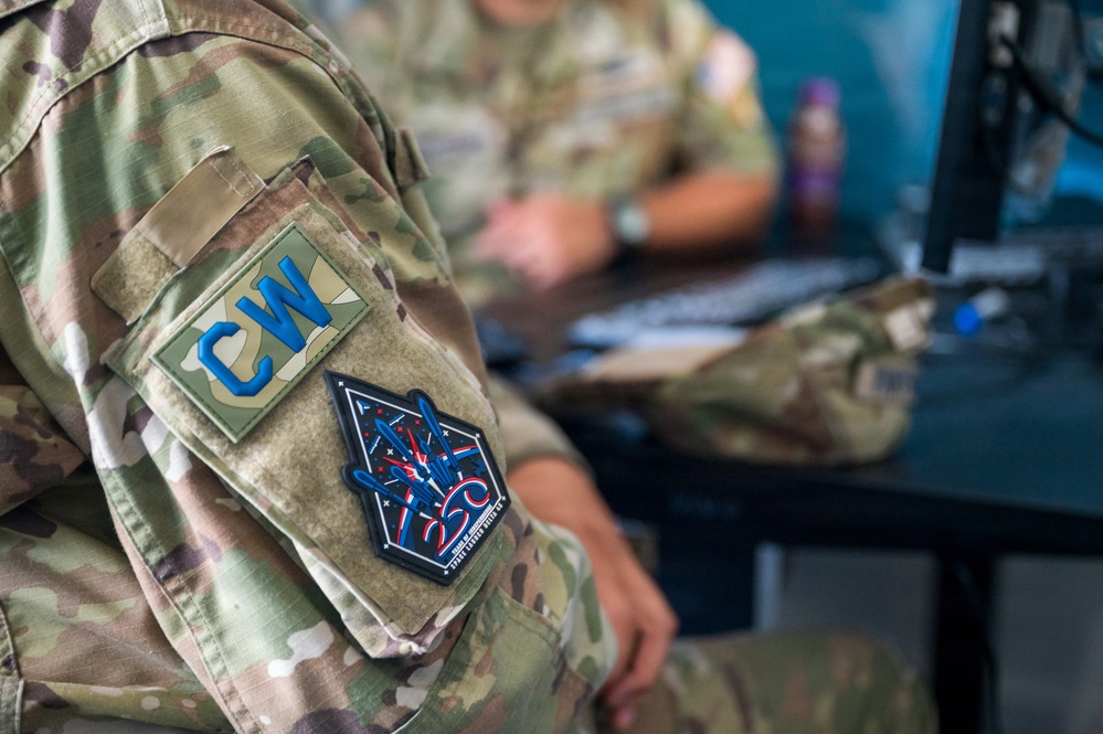 A close-up photograph of a military service member's upper arm showing two patches on an OCP (Operational Camouflage Pattern) uniform. The foreground patch reads 'CW' in blue letters on an olive/tan background, and below it is a dark pentagon-shaped morale patch with red, white, and blue design elements. A second service member is visible in the blurred background working at a computer workstation.