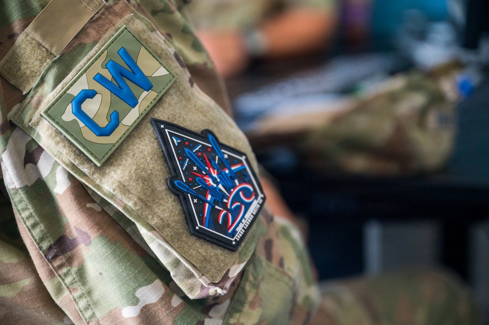 Close-up photograph of a military uniform sleeve showing two patches on a MultiCam/OCP camouflage uniform. The upper patch displays the letters 'CW' in blue on a camouflage background, while the lower patch appears to be a commemorative or unit morale patch with a dark background featuring red, white, and blue design elements and the number '25C'. The background is blurred, suggesting an indoor setting.
