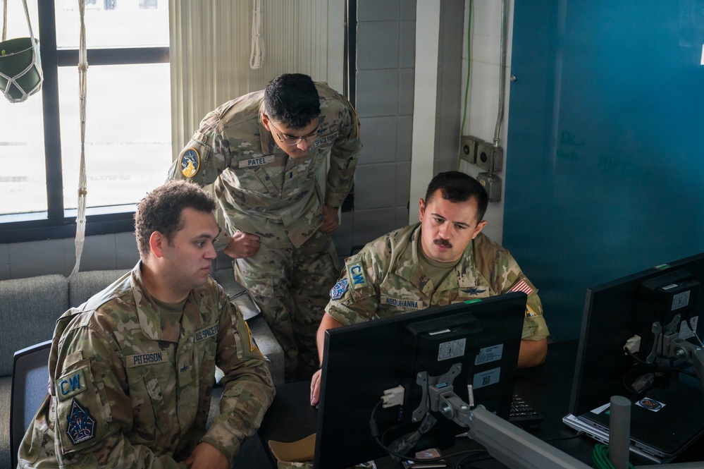Three military personnel in OCP (Operational Camouflage Pattern) uniforms are gathered around a computer monitor in what appears to be an operations or workspace environment. Name tapes visible on the uniforms read 'PETERSON', 'PATEL', and 'ABDUHANNA', with 'CW' patches and unit insignia visible. They appear to be reviewing or working on something displayed on the monitor together.
