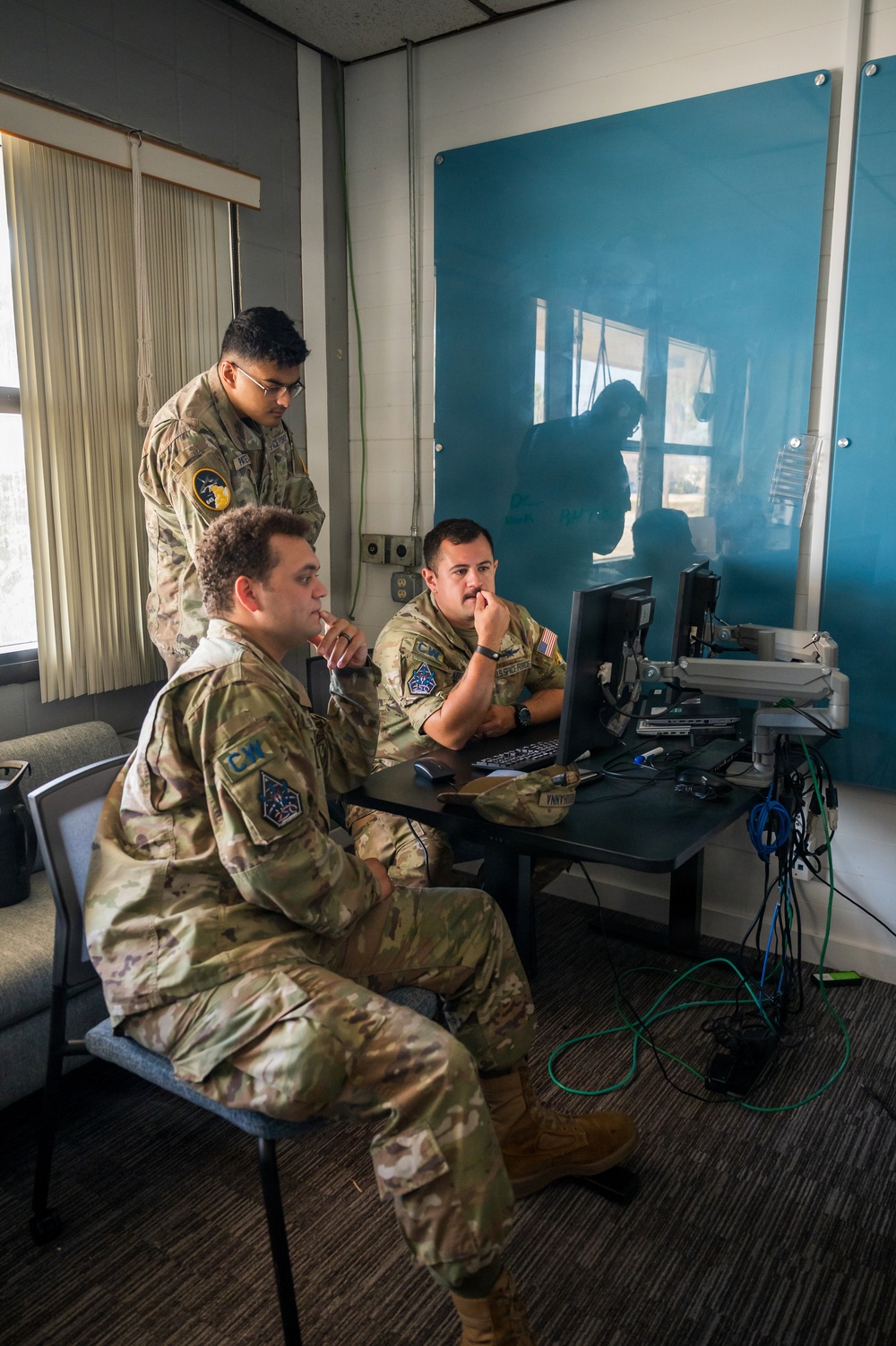 Three military personnel in OCP (Operational Camouflage Pattern) uniforms are gathered around a computer workstation in what appears to be an office or operations room. Two individuals are seated while one stands behind them, all focused on a computer monitor. The setting includes networking cables, equipment, and a blue glass board on the wall.