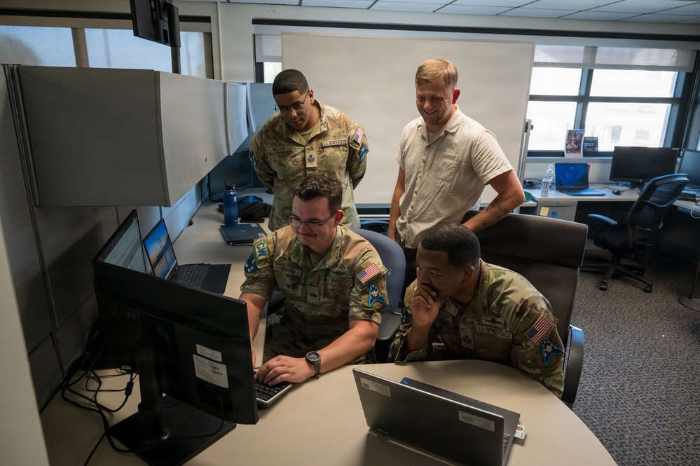 Four individuals are gathered around a computer workstation in an office environment. Three are wearing U.S. Air Force/Space Force camouflage uniforms with American flag patches, while one is in civilian attire. They appear to be collaborating on work displayed on a desktop monitor.