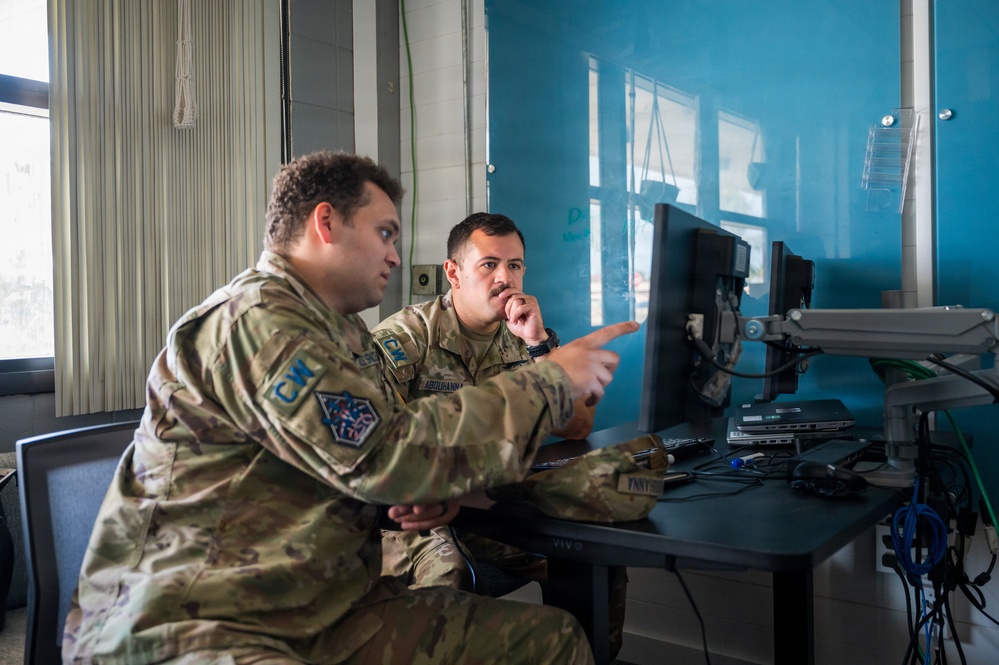 Two military personnel in OCP (Operational Camouflage Pattern) uniforms are seated at a workstation with computer monitors, appearing to review or discuss information on screen. One individual is pointing at the monitor while the other observes with a contemplative posture. The setting appears to be an office or operations center with a teal/blue wall.