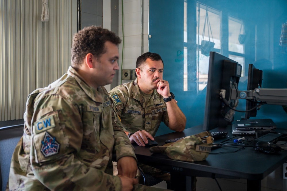 Two U.S. Space Force personnel in OCP (Operational Camouflage Pattern) uniforms are seated at a workstation reviewing content on a computer monitor. The individual on the right, whose name tape reads 'ABOUHANNA,' is leaning forward with hand raised to chin in a contemplative posture. Both appear to be engaged in a focused work or briefing session.
