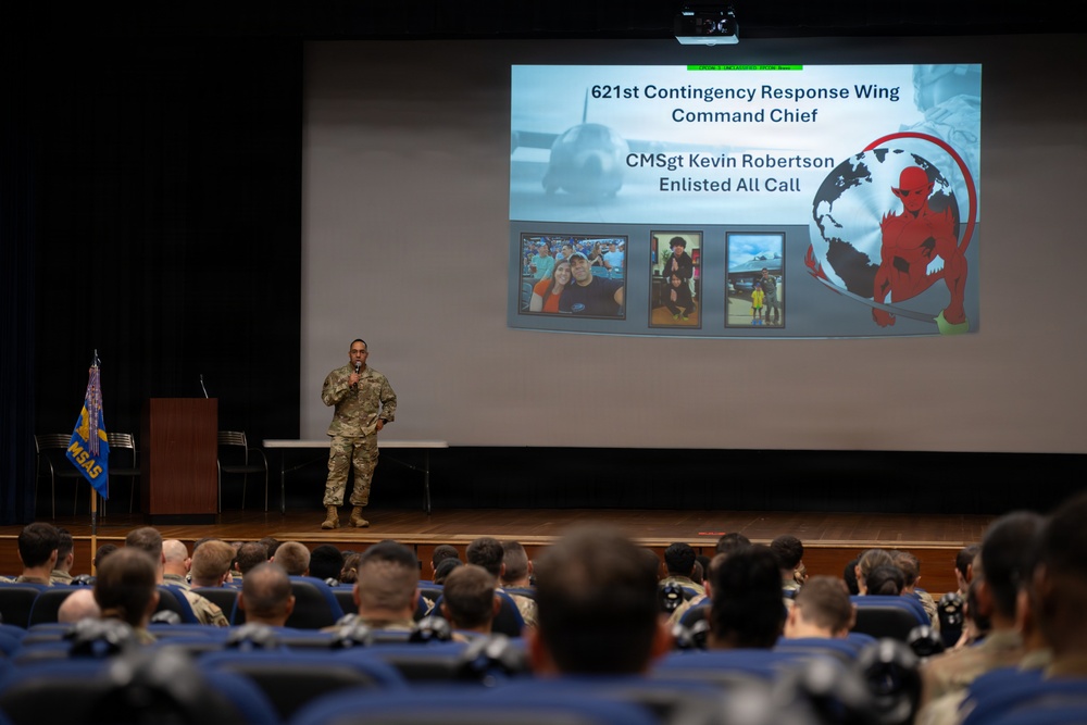 A military briefing or 'All Call' event is taking place in an auditorium, with CMSgt Kevin Robertson of the 621st Contingency Response Wing addressing a large audience of uniformed personnel. The speaker stands on a stage in front of a projected presentation slide that identifies him as the Command Chief conducting an Enlisted All Call. The audience, seated in blue chairs, consists of numerous military members in uniform.