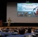 A military briefing or 'All Call' event is taking place in an auditorium, with CMSgt Kevin Robertson of the 621st Contingency Response Wing addressing a large audience of uniformed personnel. The speaker stands on a stage in front of a projected presentation slide that identifies him as the Command Chief conducting an Enlisted All Call. The audience, seated in blue chairs, consists of numerous military members in uniform.
