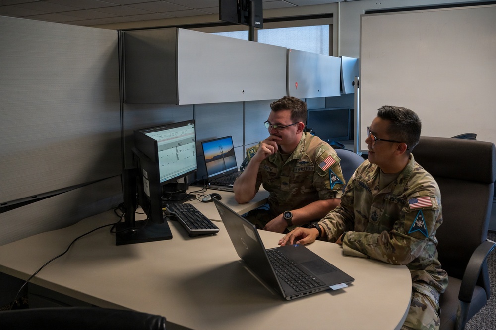 Two military personnel in OCP (Operational Camouflage Pattern) uniforms are seated at a workstation in an office cubicle environment, reviewing content on a laptop computer together. Both individuals wear U.S. flag patches and unit insignia patches on their sleeves, consistent with U.S. Air Force or Space Force uniforms. The setting appears to be a modern military administrative or operations office.
