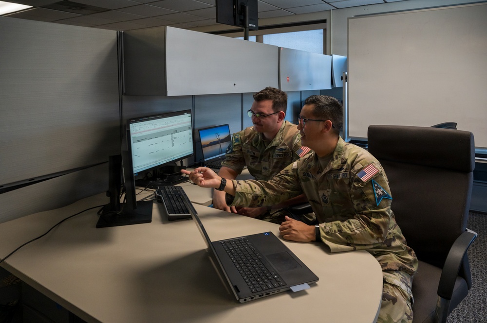 Two military personnel in U.S. Army/Space Force OCP (Operational Camouflage Pattern) uniforms are collaborating at a workstation in what appears to be an office or operations center. One individual is pointing at a desktop monitor displaying what appears to be a spreadsheet or scheduling software, while a laptop is open on the desk in the foreground. Both wear American flag patches and unit insignia on their uniforms.