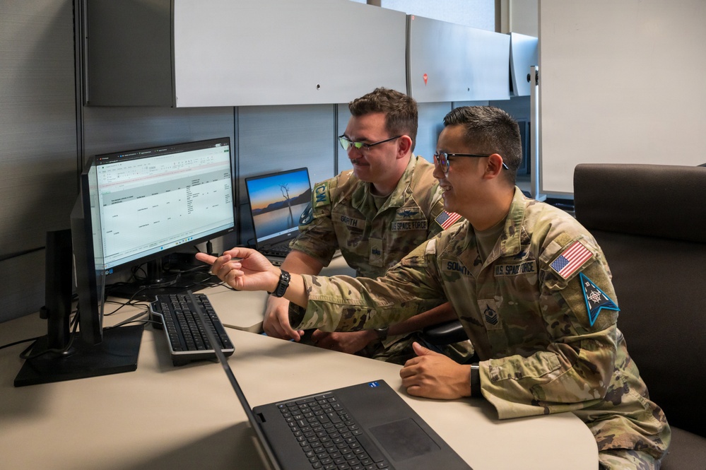 Two U.S. Space Force guardians in OCP (Operational Camouflage Pattern) uniforms are collaborating at a workstation with multiple monitors. One guardian is pointing at a screen displaying what appears to be a spreadsheet or database application while the other observes and smiles. Both wear name tapes and U.S. Space Force branch insignia patches.