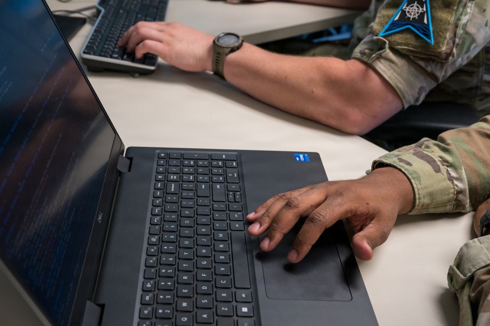Two military personnel in camouflage uniforms are working at computers in what appears to be a technical or operations environment. One individual is using a laptop in the foreground while another operates a desktop keyboard in the background. A military unit patch is visible on the sleeve of one uniform.
