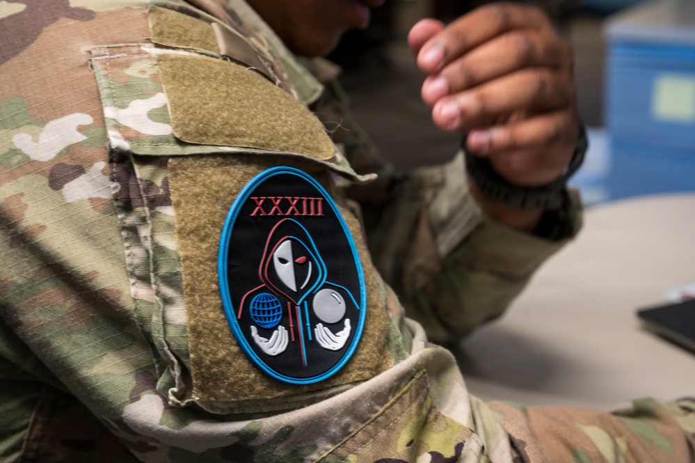 A close-up photograph of a military service member's arm wearing OCP (Operational Camouflage Pattern) uniform. A distinctive PVC morale/unit patch is prominently displayed on the upper sleeve, featuring Roman numerals 'XXXIII' and a hooded figure holding a globe and sphere. The patch has a black background with a blue oval border and red/white design elements.