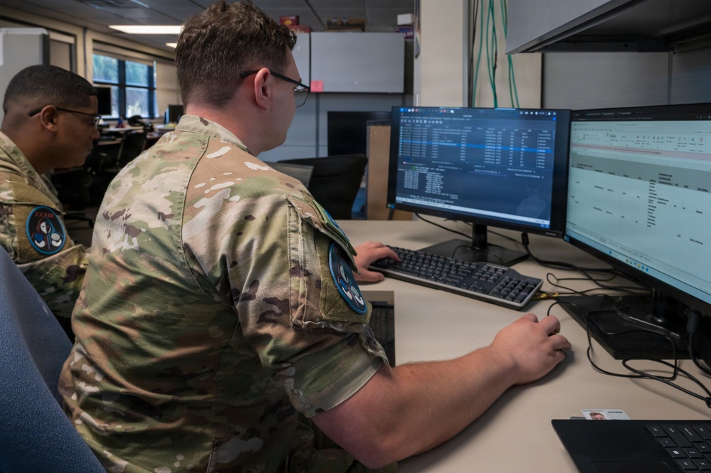 Two military personnel in OCP (Operational Camouflage Pattern) uniforms are working at a computer workstation with dual monitors in an office environment. The foreground individual is actively using a keyboard and mouse while viewing what appears to be database or logistics software on the screens. Both individuals wear unit patches on their sleeves, suggesting an Air Force or Space Force assignment.