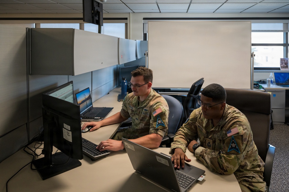 Two military personnel in OCP (Operational Camouflage Pattern) uniforms are working at a shared desk in what appears to be an office or operations center. Both individuals are focused on laptop computers, with additional monitors visible on the desk. Their uniforms display U.S. flag patches and what appear to be U.S. Space Force delta insignia patches.