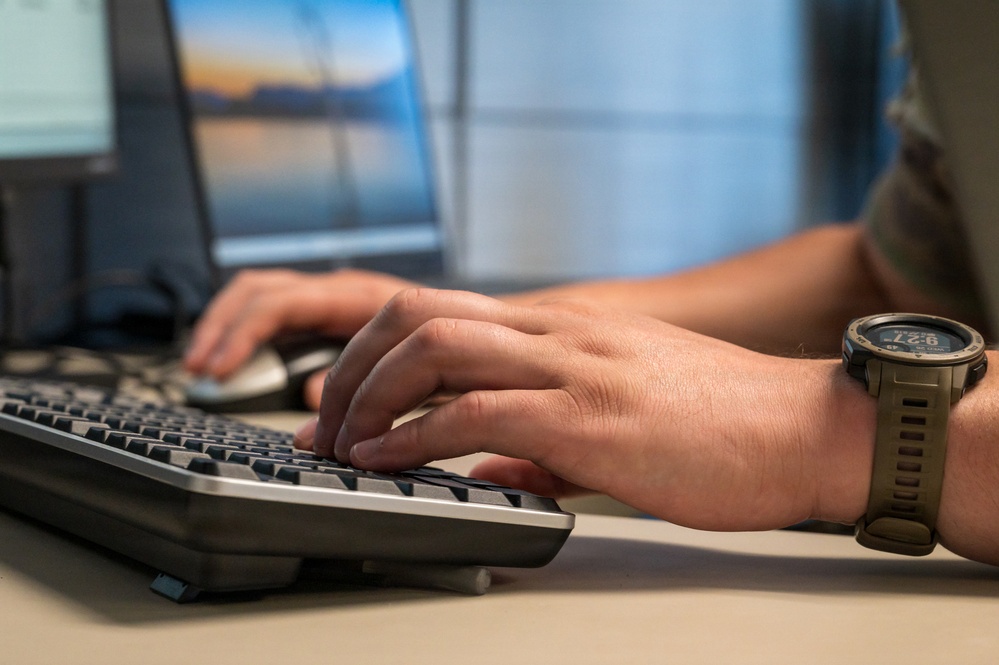 A person is typing on a desktop keyboard while simultaneously using a computer mouse. The close-up shot focuses on both hands positioned over a dark ergonomic keyboard, with a tactical-style wristwatch visible on the right wrist. A blurred monitor displaying what appears to be a landscape wallpaper is visible in the background.