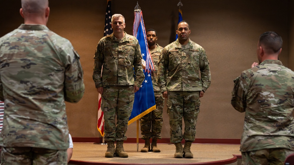 A formal military ceremony is taking place in an indoor venue, likely a change of command or award ceremony. Several service members in OCP (Operational Camouflage Pattern) uniforms are positioned on and around a raised platform, flanked by the U.S. flag and what appears to be a U.S. Air Force organizational flag. Two individuals face the platform from the foreground, suggesting a formal presentation or review.