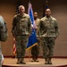 A formal military ceremony is taking place in an indoor venue, likely a change of command or award ceremony. Several service members in OCP (Operational Camouflage Pattern) uniforms are positioned on and around a raised platform, flanked by the U.S. flag and what appears to be a U.S. Air Force organizational flag. Two individuals face the platform from the foreground, suggesting a formal presentation or review.