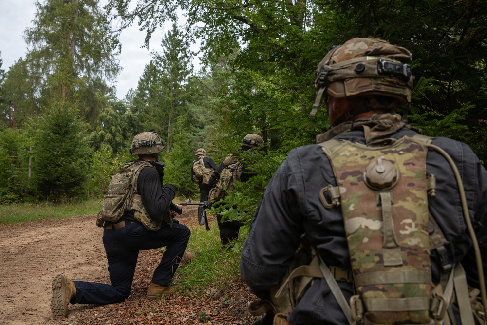The image shows a group of military personnel conducting what appears to be a tactical field exercise in a forested environment. Soldiers in camouflage gear and helmets are moving along a dirt trail at the edge of a tree line, with at least one soldier in a kneeling position. The scene is consistent with a training exercise or patrol operation.