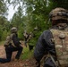 The image shows a group of military personnel conducting what appears to be a tactical field exercise in a forested environment. Soldiers in camouflage gear and helmets are moving along a dirt trail at the edge of a tree line, with at least one soldier in a kneeling position. The scene is consistent with a training exercise or patrol operation.
