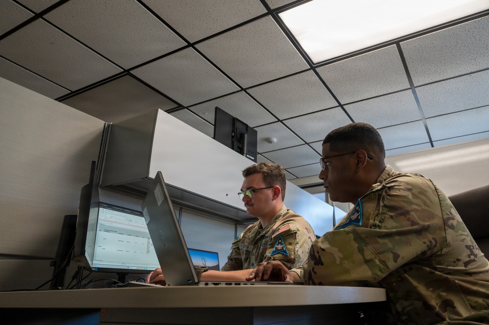 Two military personnel in camouflage uniforms are working together at a computer workstation in what appears to be an office or operations center. One individual is typing on a laptop while the other observes the screen. The setting suggests a collaborative work or training environment within a military facility.