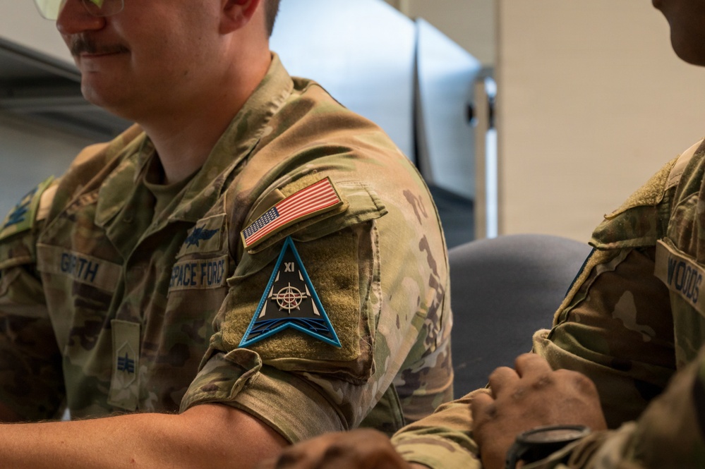 The image shows a U.S. Space Force service member in OCP (Operational Camouflage Pattern) uniform seated alongside other military personnel. The central figure displays a Space Force branch tape and a distinctive triangular unit patch with 'XI' designation on the sleeve. The setting appears to be an indoor military briefing or meeting environment.