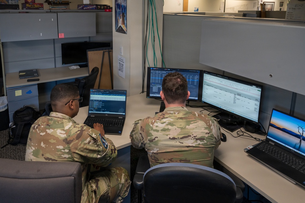 Two military personnel in OCP (Operational Camouflage Pattern) uniforms are seated at a workstation in an office environment, working on multiple computers. The individual on the left is using a laptop displaying what appears to be a command-line interface or code, while the person on the right works with dual monitors showing spreadsheet or database applications. The setting appears to be a military office or operations center with cubicle-style partitions.