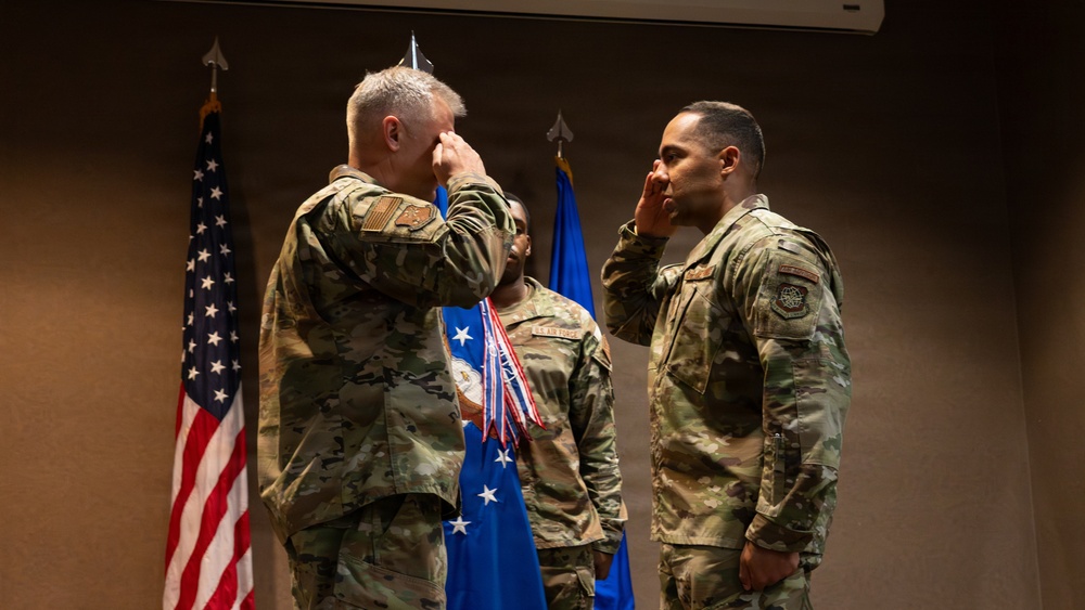Two military personnel in OCP (Operational Camouflage Pattern) uniforms are exchanging salutes during what appears to be a formal military ceremony. A third service member stands in the background holding what appear to be medals or ribbons. The setting includes a U.S. flag and a blue Air Force flag, suggesting this is a U.S. Air Force recognition or awards ceremony.