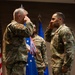 Two military personnel in OCP (Operational Camouflage Pattern) uniforms are exchanging salutes during what appears to be a formal military ceremony. A third service member stands in the background holding what appear to be medals or ribbons. The setting includes a U.S. flag and a blue Air Force flag, suggesting this is a U.S. Air Force recognition or awards ceremony.