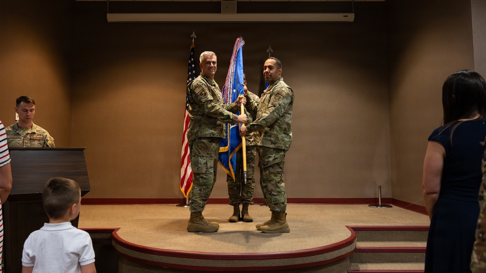 A military change of command ceremony is taking place indoors, with two uniformed service members jointly holding a blue guidon flag on a raised stage. The ceremony appears to be a formal transfer of command, a traditional military ritual. Observers including a child and a civilian woman are visible in the foreground.