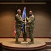 A military change of command ceremony is taking place indoors, with two uniformed service members jointly holding a blue guidon flag on a raised stage. The ceremony appears to be a formal transfer of command, a traditional military ritual. Observers including a child and a civilian woman are visible in the foreground.